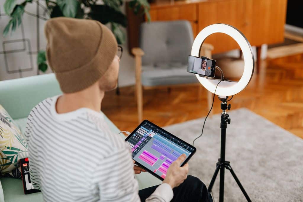 YouTube Equipment Ring Light. A man wearing a brown beanie hat holds an iPad and speaks into a smart phone on a ring light tripod. 